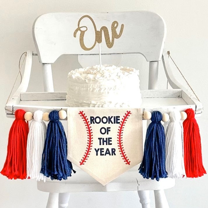 Rookie of the year baseball banner with navy, white and red tassels and wooden beads attached to a highchair that has a cake with a one cake topper.