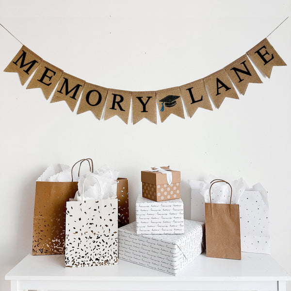 Memory Lane banner made from burlap. The wording is in black capital letters and there is a painted image of a black graduation cap with a blue tassel between the words. The flags are stitched at the top and there is string going through to hang.