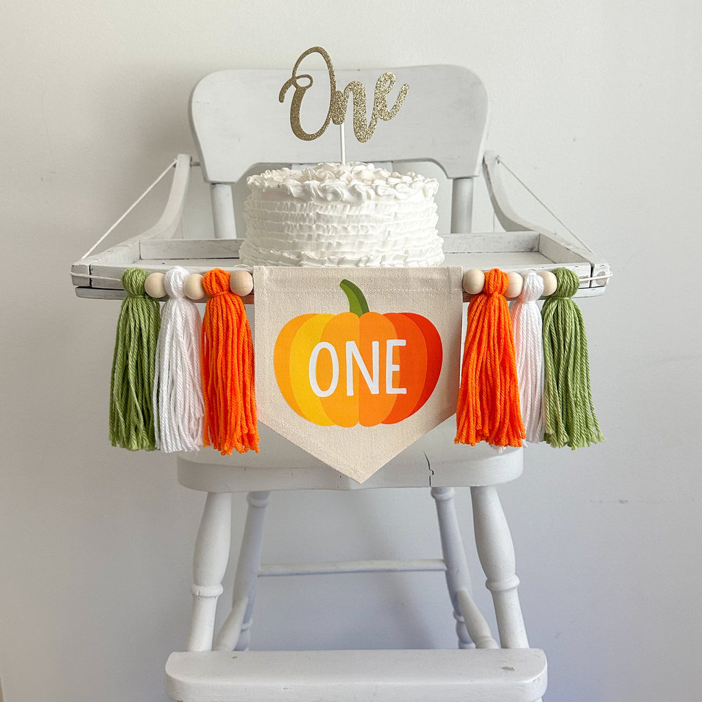 A canvas center flag with an ombre style pumpkin on it in orange with a green stem and the word ONE in white in the middle of the pumpkin. There is an orange, white and sage green tassel on either side of the center flag separated by 6 wooden beads.