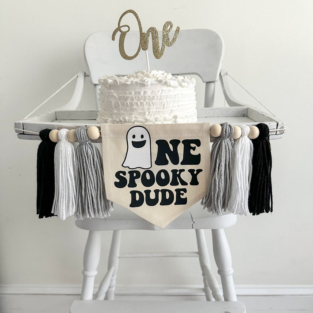 A canvas flag measuring 5x7 inches with a white ghost followed by the letters NE in black. The words SPOOKY DUDE are below that. There is a gray, white and black tassel on each side each separated by a wooden bead, for a total of 6 tassels and beads.