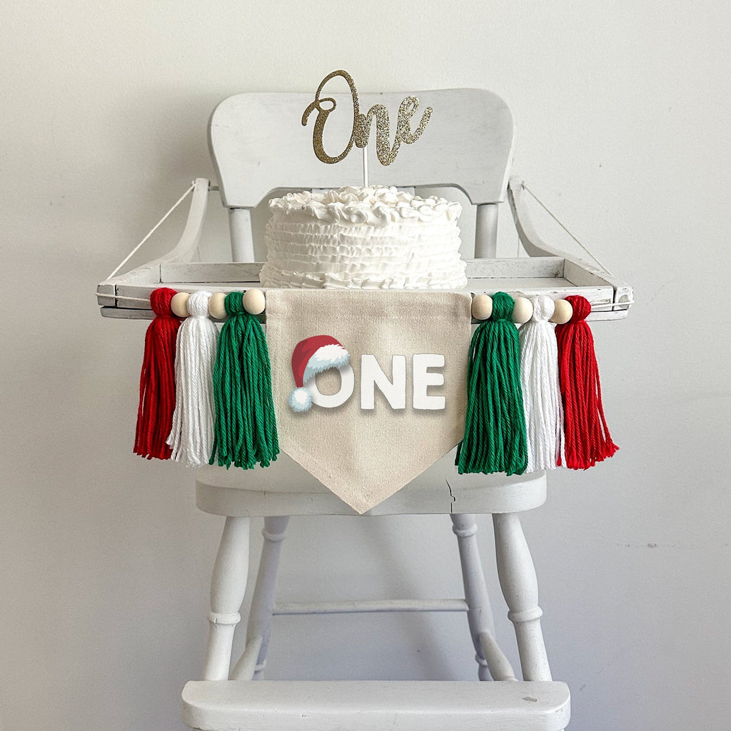 A 5x7 canvas flag with the word ONE in white with a shadow and a red Santa hat sitting atop the letter O. There are 6 tassels, 3 on each side in green, white and red. They are each separated with a wooden bead for a total of 6 beads.