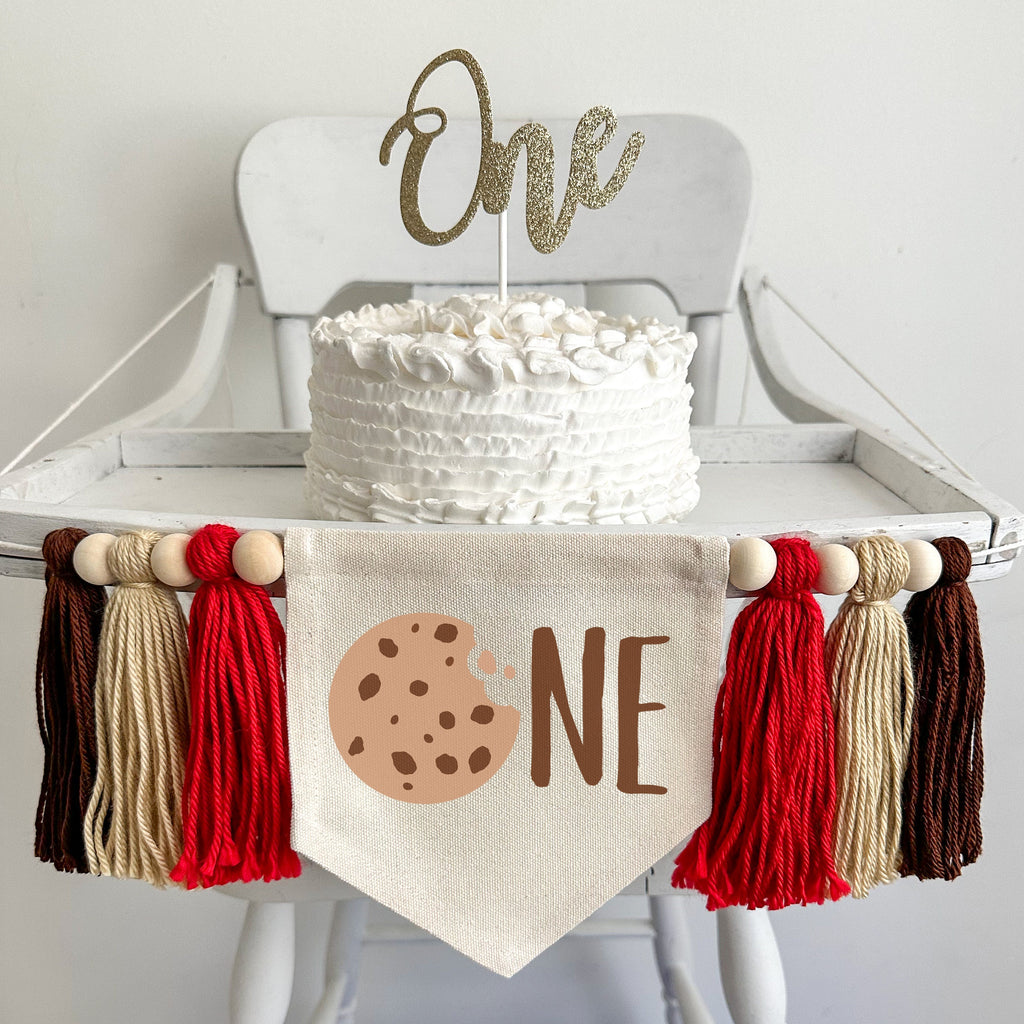 A 5 by 7 canvas flag with a chocolate chip cookie followed by the letters NE in brown to spell the word ONE. On each side there is a red, tan and brown tassel, each separated by a wooden bead for a total of 6 tassels and 6 beads.