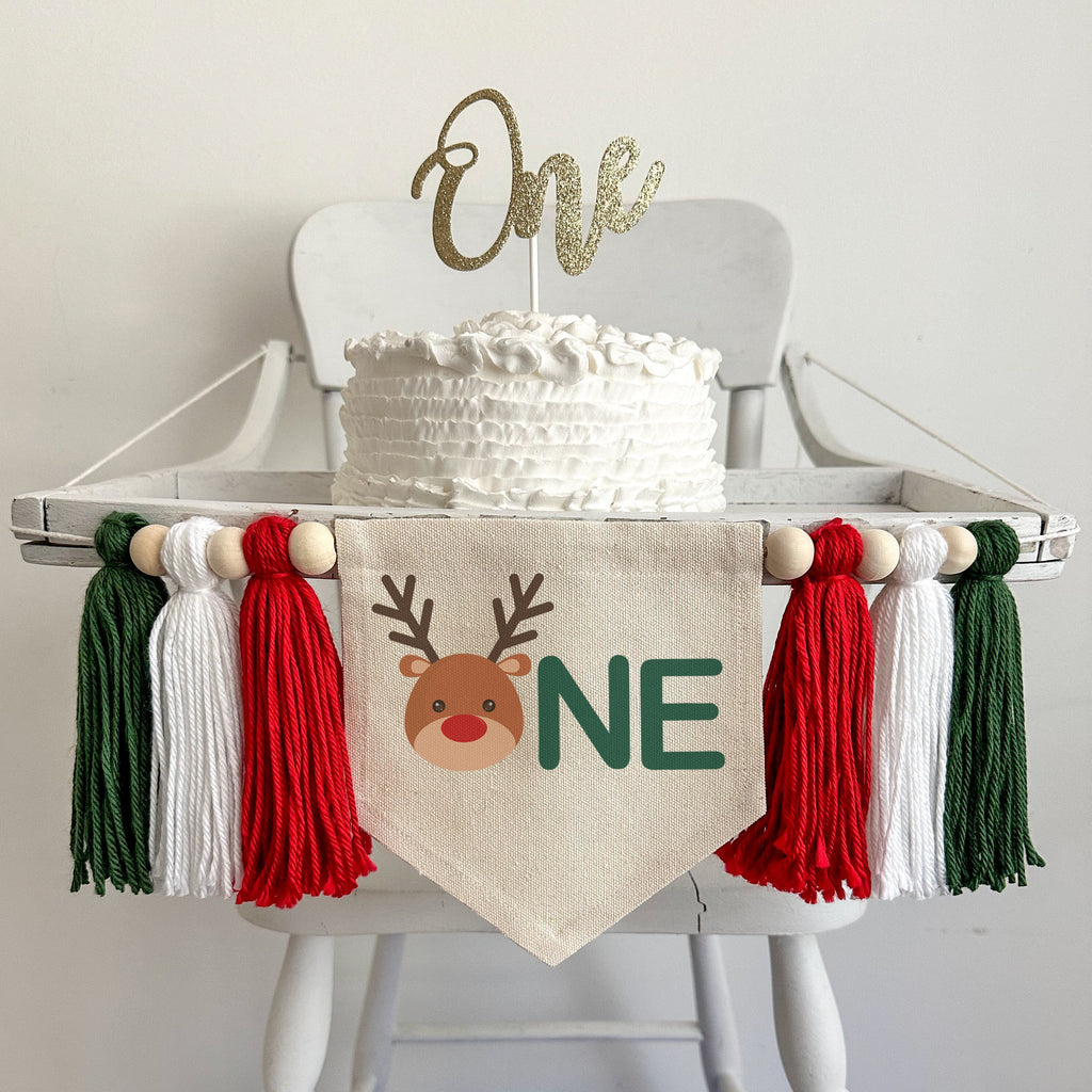 A 5 x 7 canvas center flag with a cute reindeer head to make the O followed by the letters NE in green which makes the word ONE. There is a red, white and dark green 5 inch yarn tassel on each side, each separated by a wooden bead.