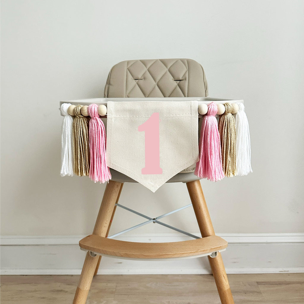 A 5 x 7 inch canvas flag with a pink number 1 on it. On each side is a pink, tan and white colored 5 inch yarn tassel each separated by a wooden bead. There is a total of 6 tassels and 6 wooden beads.