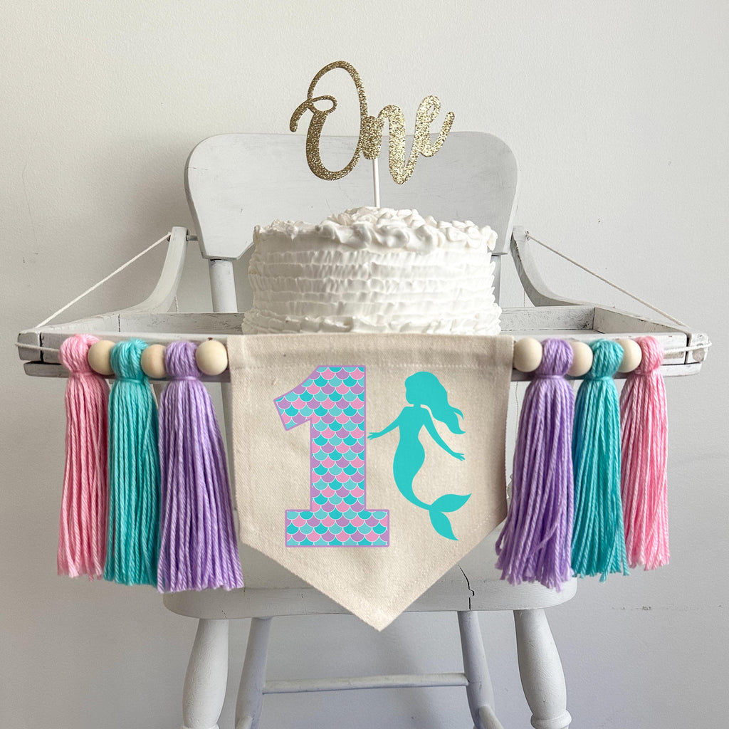 A 6 by 7 inch canvas center flag with a large number 1 with pink, lavender, and light aqua scales with an aqua mermaid silhouette beside it. On each side there is a 5 inch lavender, light aqua and pink yarn tassel each separated by a wooden bead.