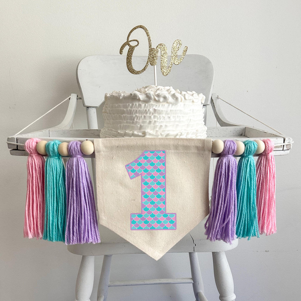 A 6 by 7 inch canvas center flag with a number one outlined in lavender with pink, lavender and light aqua mermaid scales on it. On each side there is a 5 inch lavender, light aqua and pink yarn tassel each separated by a wooden bead.