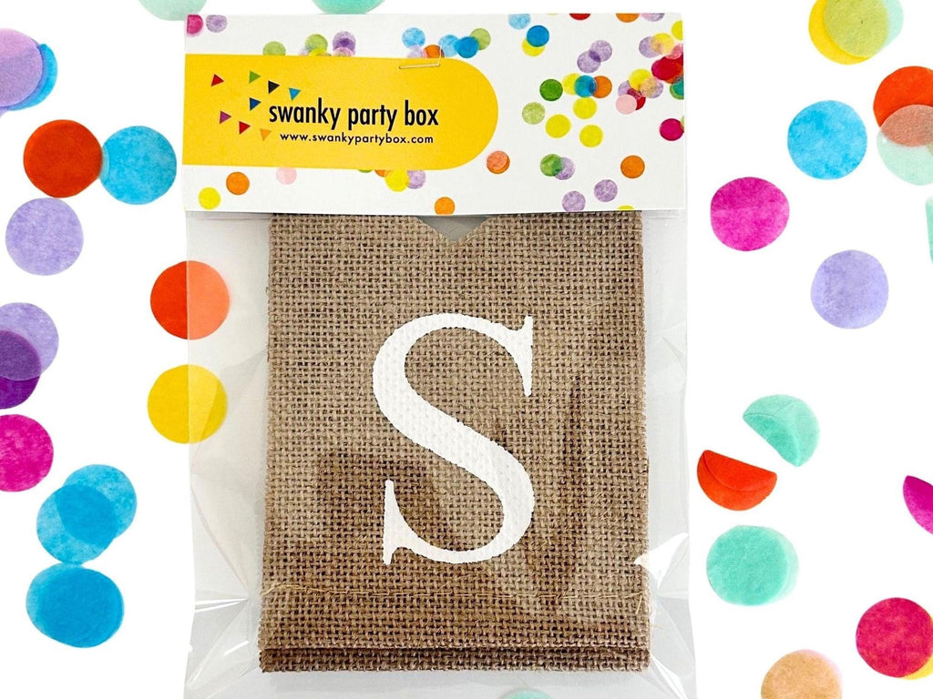 A burlap banner with a large white letter "S" on it, placed in a clear plastic bag. The banner is surrounded by colorful confetti.