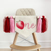 A canvas high chair banner featuring a red strawberry as the letter O followed by an ne spelling out the word "one" on it, along with tassels in pink and red.