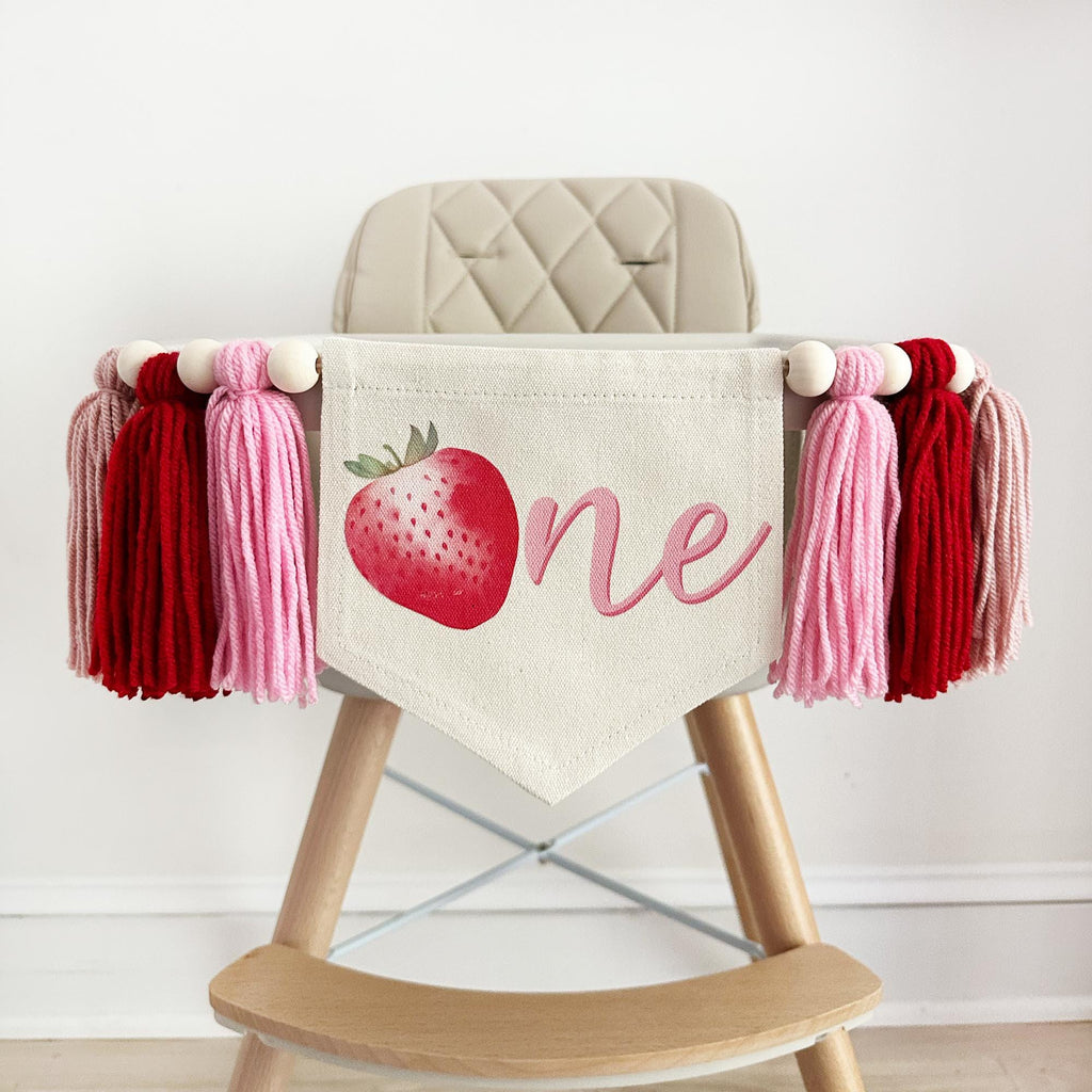 A canvas high chair banner featuring a red strawberry as the letter O followed by an ne spelling out the word "one" on it, along with tassels in pink and red.