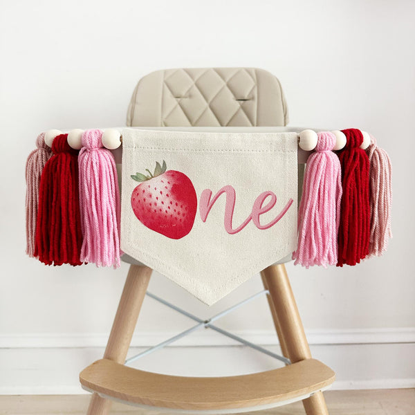 A canvas high chair banner featuring a red strawberry as the letter O followed by an ne spelling out the word "one" on it, along with tassels in pink and red.