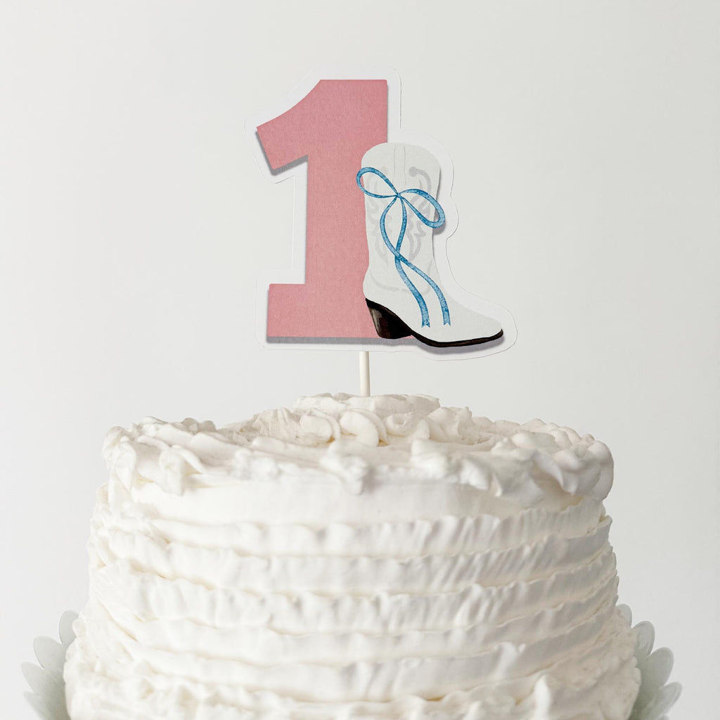 A cake with a pink number one topper featuring a white boot with a blue ribbon.