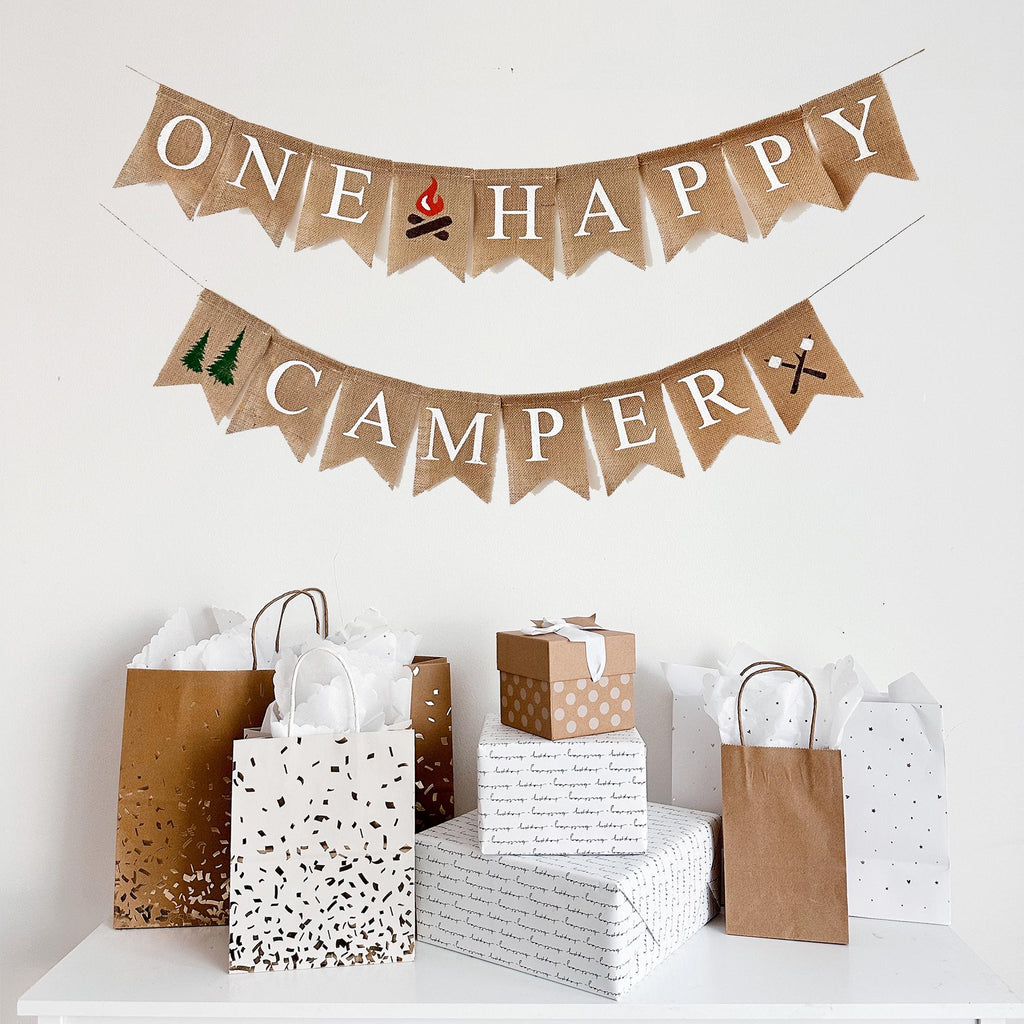 A burlap banner that reads "ONE HAPPY CAMPER" and various gift bags and boxes arranged below it.
