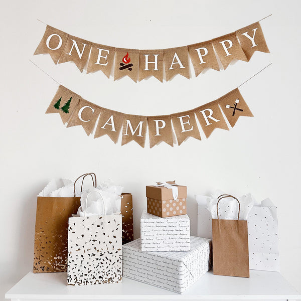 A burlap banner that reads "ONE HAPPY CAMPER" and various gift bags and boxes arranged below it.