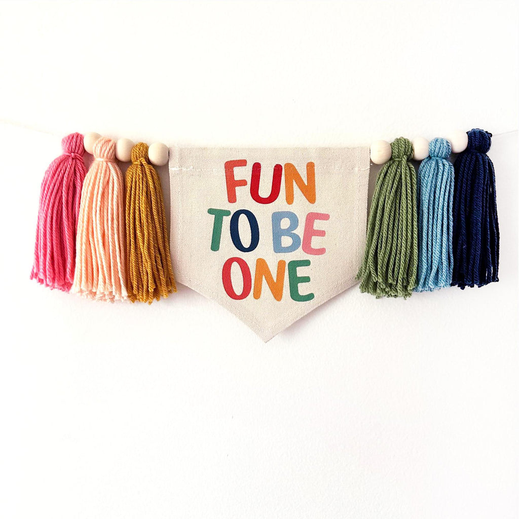 A banner with the text "FUN TO BE ONE" is hanging on a white wall, surrounded by colorful tassels in various shades of pink, orange, yellow, green, blue, and navy.