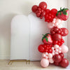 A large balloon arch made of red and pink balloons, including some that appear to be strawberries, is displayed against a white wall.