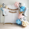 a festive scene with a banner that reads "a little cowgirl is on the way" and a variety of balloons in different colors, including white, pink, blue, and black and white. There are also two large white boots hanging on the wall, and the entire setup is placed on a wooden floor.