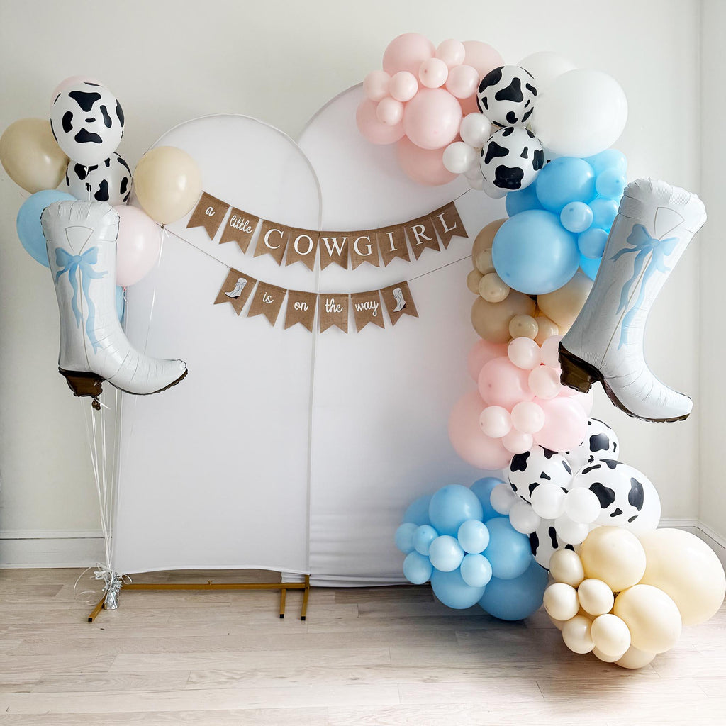 a festive scene with a banner that reads "a little cowgirl is on the way" and a variety of balloons in different colors, including white, pink, blue, and black and white. There are also two large white boots hanging on the wall, and the entire setup is placed on a wooden floor.