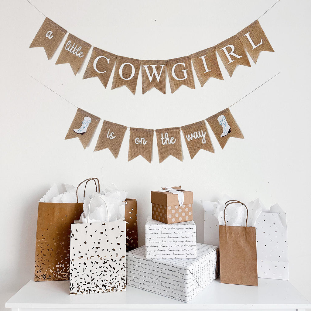 a festive scene with a banner that reads "a little cowgirl is on the way" and several gift bags and boxes arranged on a white surface.