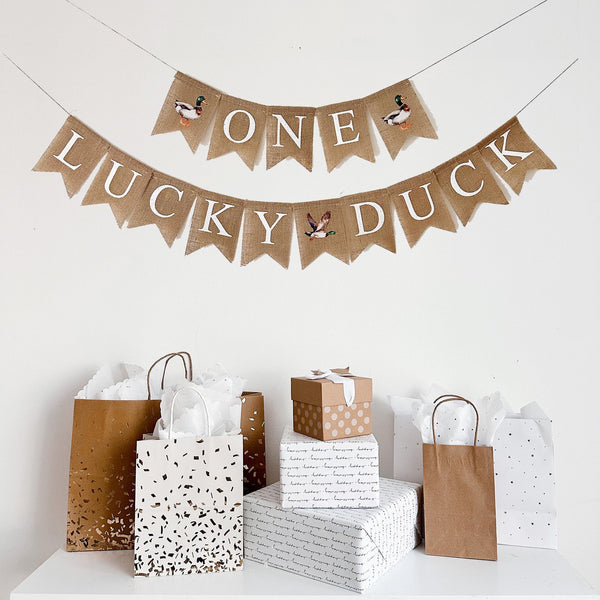 a festive scene with a banner that says "Lucky Duck" and several gift bags and boxes arranged on a white surface.