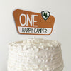 A white cake with a brown topper that says "ONE HAPPY CAMPER".