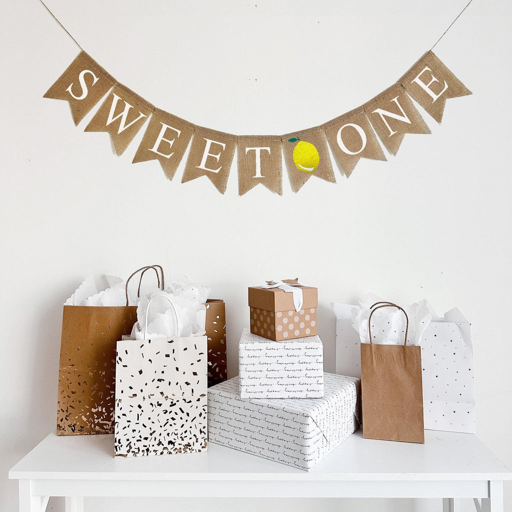 a banner with the word "SWEET ONE" with an image of a lemon between the words, hanging above a table with gifts