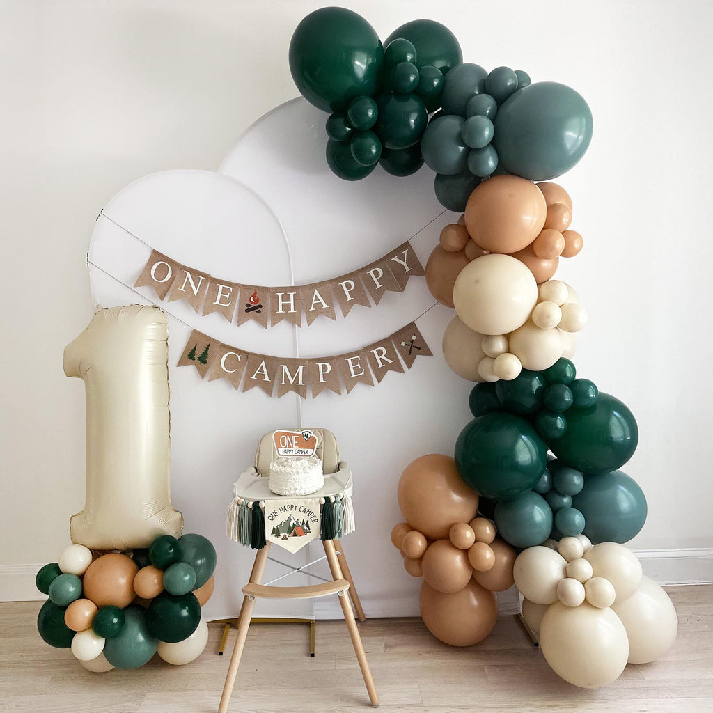 A festive one-year-old's birthday party setup, featuring a white backdrop, a banner that reads "ONE HAPPY CAMPER", and a high chair with a cake on it. The centerpiece is a large balloon arch made of various colored balloons, including green, brown, and beige.