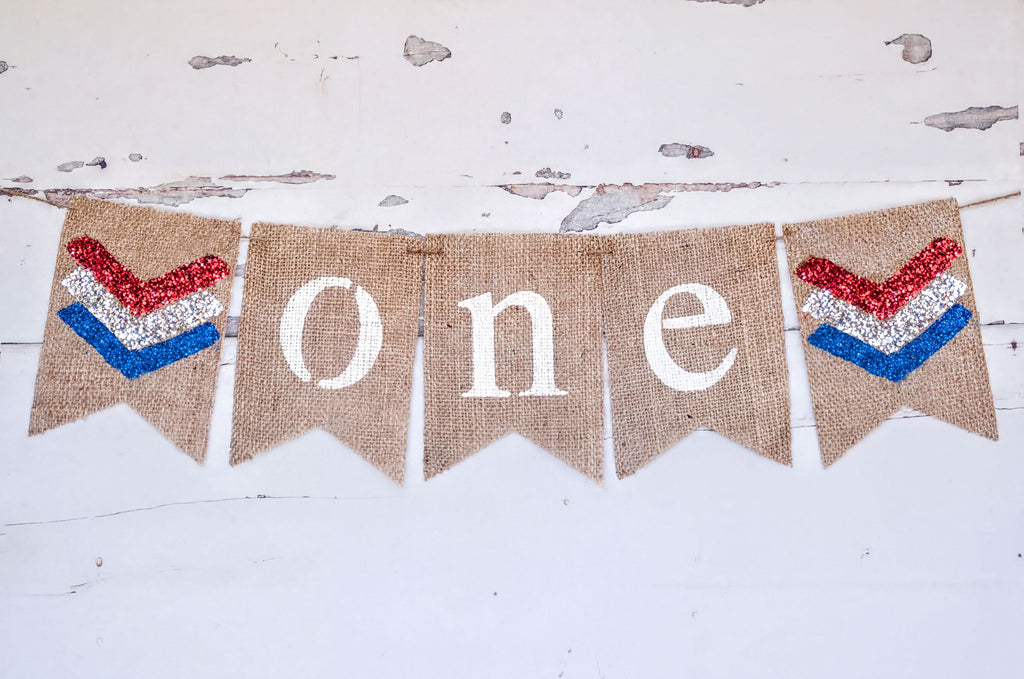 A banner with the word "ONE" written on it, made of burlap and decorated with patriotic colors.