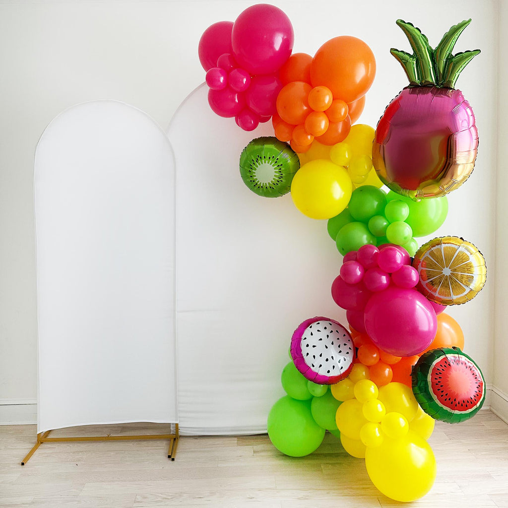 A colorful balloon arrangement decorated with different fruit balloons, including a pineapple, kiwi, and other assorted fruits, is displayed against a white wall.