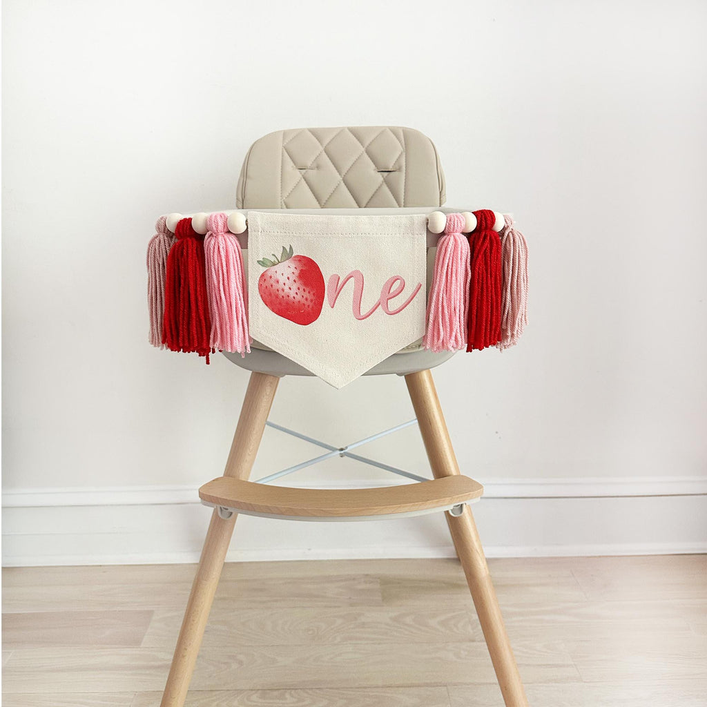 A canvas high chair banner featuring a red strawberry as the letter O followed by an "ne" spelling out the word "one" on it, along with tassels in pink and red.