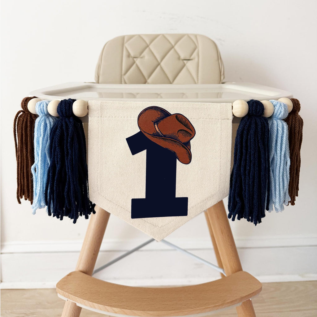 A beige high chair with a white cushion and a banner featuring a cowboy hat and the number 1 printed on it.