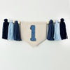 This image shows a tassel banner hanging on the wall with dark blue and light blue tassels and a banner that displays the number 1 in a denim print.