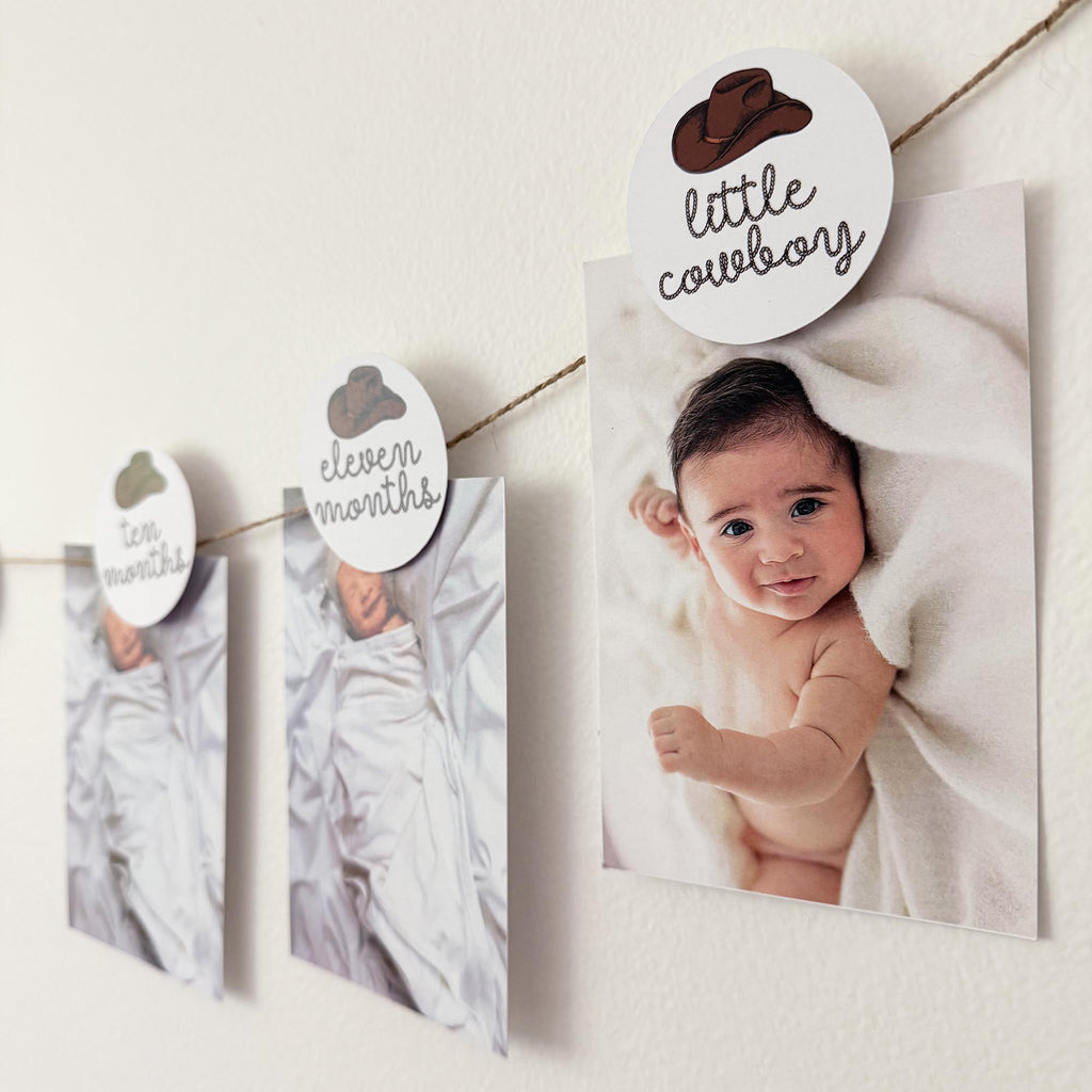 A baby photo is displayed on a wall, with a string of small paper clips hanging from it, each attached to a larger paper clip that has a picture of a baby and the text "little cowboy" written on it.