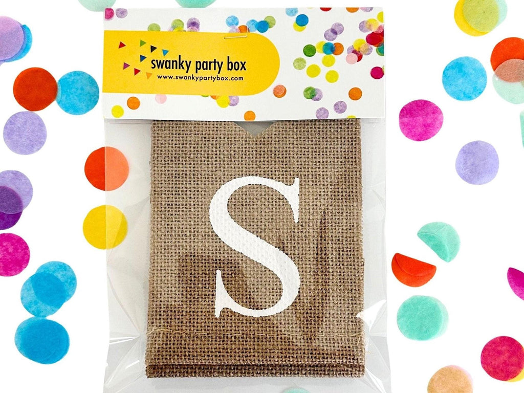A burlap banner in a clear plastic bag. The bag has a colorful bag topper with the Swanky Party Box logo and a pattern of colorful confetti.