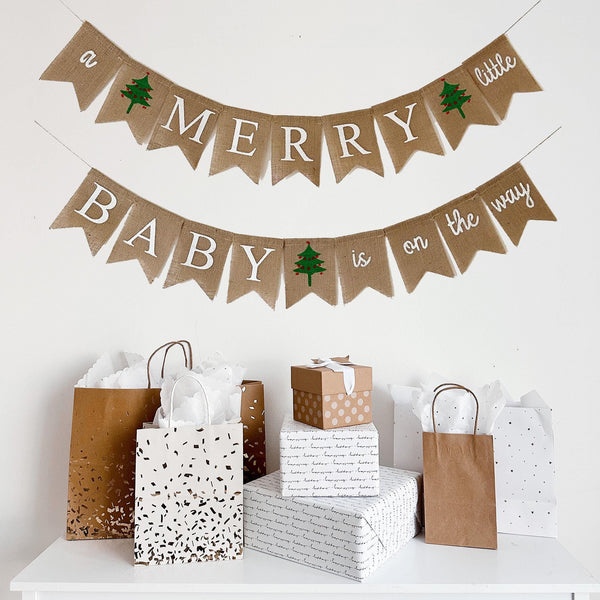 a festive scene with a banner that reads "a Merry Baby is on the way" and several gift bags and boxes arranged on a white surface.