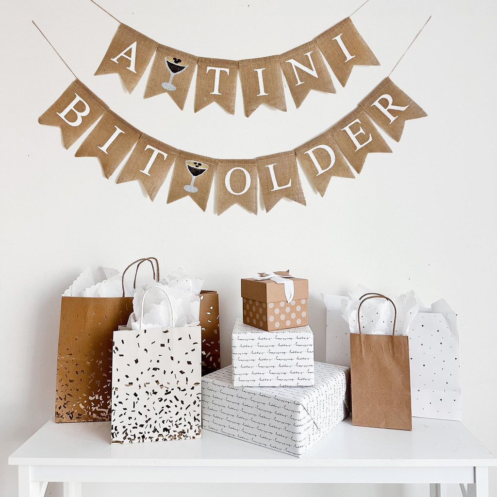 a banner that reads "A TINI BIT OLDER" with expresso martini images between the words, the banner is hanging above a table with various gift bags and boxes.