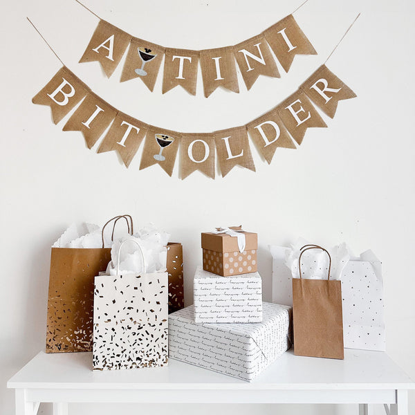 a banner that reads "A TINI BIT OLDER" with expresso martini images between the words, the banner is hanging above a table with various gift bags and boxes.