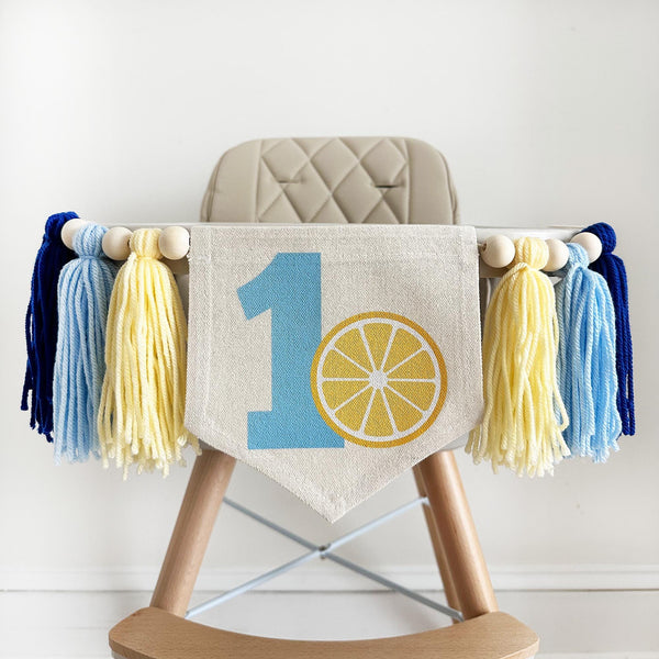 A high chair banner featuring a blue number one and a yellow lemon slice surrounded by yarn tassels in dark blue, light blue and pale yellow.