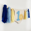 A banner with tassels hanging on a wall, featuring a blue number one and a yellow lemon slice surrounded by yarn tassels in dark blue, light blue and pale yellow.
