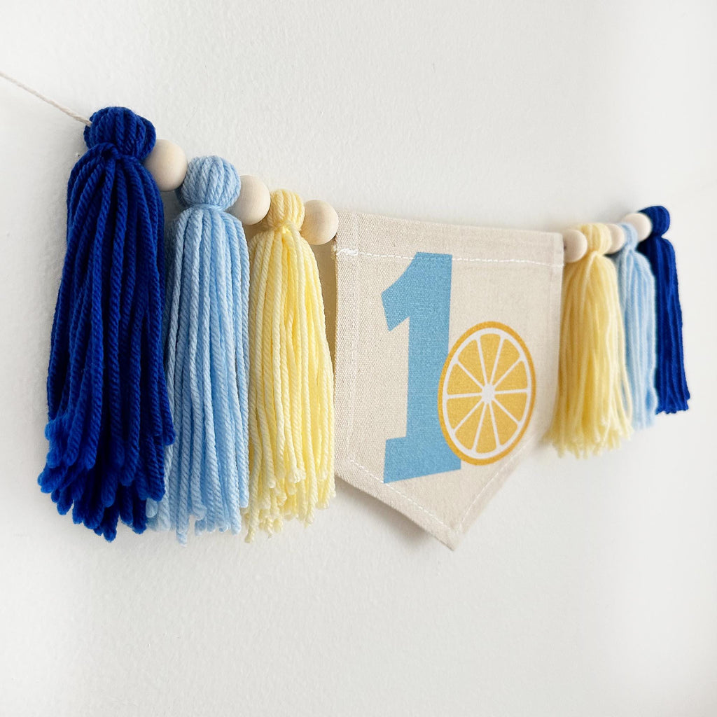 A banner with tassels hanging on a wall, featuring a blue number one and a yellow lemon slice surrounded by yarn tassels in dark blue, light blue and pale yellow.