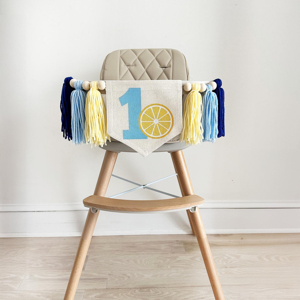 A high chair with a banner displaying the number 1 and a lemon graphic, along with blue and yellow tassels.