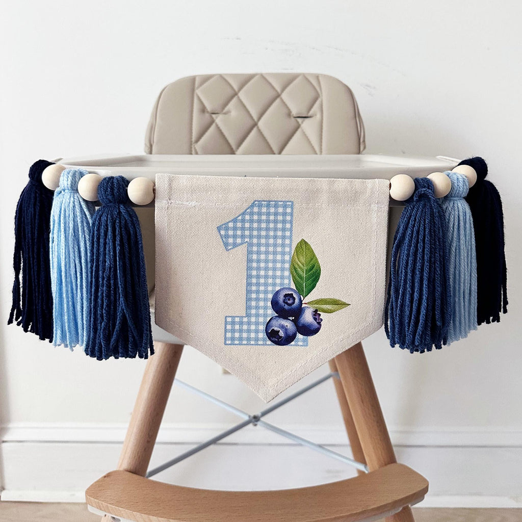 A high chair banner displaying a blue gingham printed number 1 and blueberries hanging from the side.