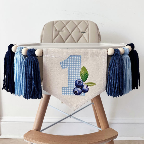 A high chair banner displaying a blue gingham printed number 1 and blueberries hanging from the side.