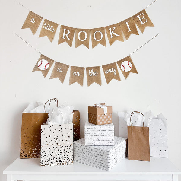 a banner with the text "a little ROOKIE is on the way" and baseball images at the end of the lower banner hanging above a table with a collection of gift bags and boxes.
