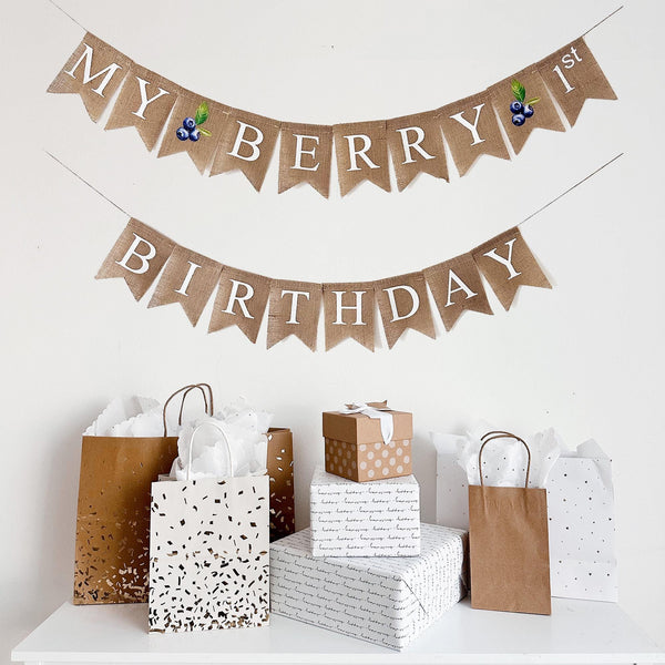 a birthday celebration setup with a banner that reads "My Berry 1st Birthday" and various gift bags and boxes arranged below it.
