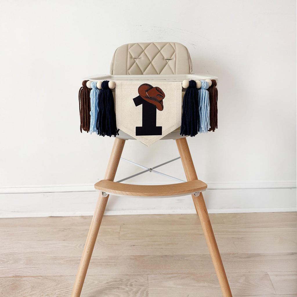 A beige high chair with a tasseled banner featuring a number one and a cowboy hat design, placed on a wooden floor against a white wall.