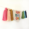 A banner with colorful tassels hanging on a white wall, and a sign with the text "FUN TO BE ONE" in the center.