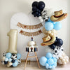 a festive scene with a cake, balloons, and a banner that reads "My First Rodeo". The cake is placed on a table, surrounded by a large number one balloon and several smaller balloons in various colors, including blue, white, and black. The overall setting appears to be a celebration for a first birthday, with the balloons and decorations creating a cheerful atmosphere.