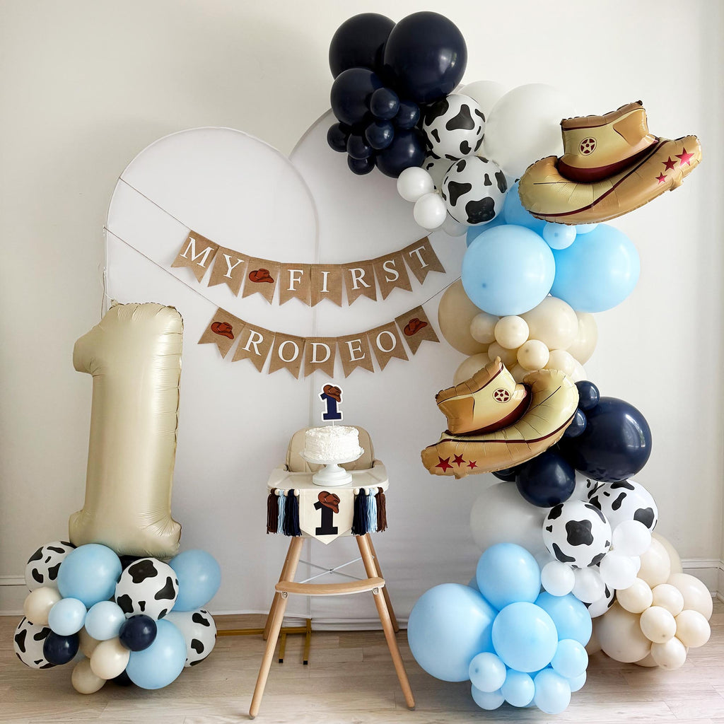 a festive scene with a cake, balloons, and a banner that reads "My First Rodeo". The cake is placed on a table, surrounded by a large number one balloon and several smaller balloons in various colors, including blue, white, and black. The overall setting appears to be a celebration for a first birthday, with the balloons and decorations creating a cheerful atmosphere.