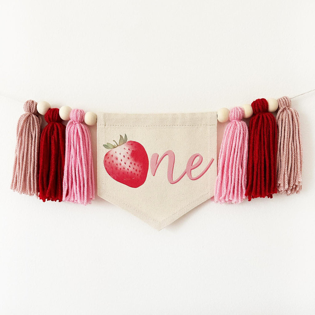 A canvas high chair banner hanging on a wall featuring a red strawberry as the letter O followed by an ne spelling out the word "one" on it, along with tassels in pink and red.