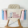 A beige high chair with a banner hanging from it that says "MY FIRST RODEO" and features a graphic of a cowboy boot framed by light pink, light blue and white yarn tassels.